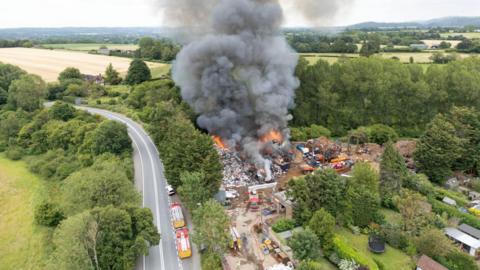 Drone shot of flames and black smoke coming from John Huntley scrapyard. Fire vehicles can be scene on the site and an adjoining road. The site is surrounded by fields, trees and private gardens.