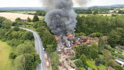 Big Ladder Photography Drone shot of flames and black smoke coming from John Huntley scrapyard. Fire vehicles can be scene on the site and an adjoining road. The site is surrounded by fields, trees and private gardens.
