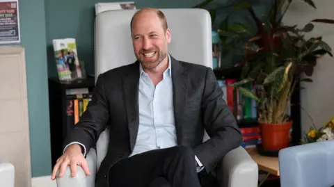 PA Medios Príncipe William sonriendo a la cámara y sentado en un sillón. Lleva un traje oscuro y una camisa pálida.