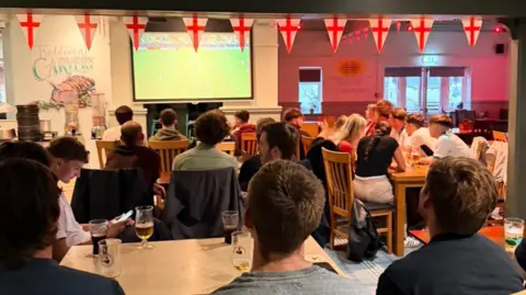 Emma Davis A crowded bar in Saltash with people all looking at the TV where the England vs Mexico game is showing. There is England bunting hanging on the wall. 