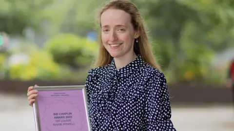 Louise Powell smiling into the camera while holding a purple certificate in a silver frame. The certificate reads: 'Sid Chaplin Award 2023 Dr Louise Marie Powell'. Powell has blonde, long hair, the front of which is tied back. She is wearing a blue and white blouse with spots on it and is standing outside in front of greenery which is out of focus.