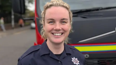 Ciara McGurk has curly blonde hair and is wearing a NIFRS navy uniform.
She is smiling and standing in front of a fire engine.