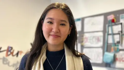 Hannah Reid, smiling at the camera. She is wearing a blue top with a white scarf and is standing in front of a noticeboard.