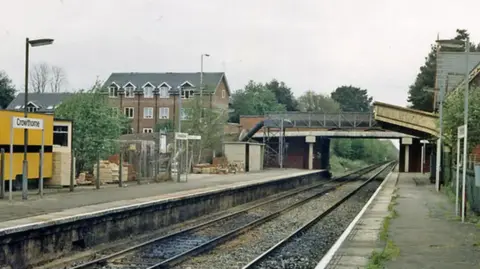 View of a railway station with two parallel train tracks running into the distance. The platforms are separated by the tracks, and a footbridge spans across them near the center of the image. A yellow portable building with a visible station sign reading “Crowthorne” stands on the left platform. Residential buildings and trees are visible in the background. The sky is overcast, and the platforms appear quiet with no trains or passengers present.