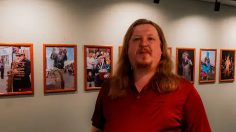 Simon Shaw Adam Parkin is standing in front of a row of his photographs. He has long, brown hair, facial hair and is wearing a red polo shirt. He is smiling slightly at the camera. On of the images over his left shoulder depicts a man wearing a black and gold uniform while playing the saxophone.