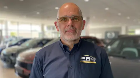 Robert is wearing glasses and a branded blue shirt stands inside a car showroom, with multiple vehicles blurred in the background under bright indoor lighting and wide windows visible behind slightly.