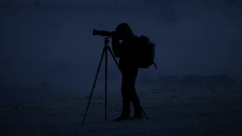 Steve Geliot A man completely covered in shadow stands taking a picture on a big camera on an icy morning. 