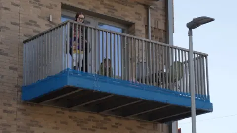 Woman stands alone on a first-floor balcony alongside her children playing with a blue safety covering at the base.