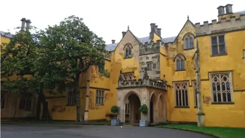 The yellow-tinted stone exterior of Ashton Court Mansion with many Gothic-style windows over three floors, crenellated ramparts and a grand entrance that has several archways. Two green trees stand close to the mansion, to the left 