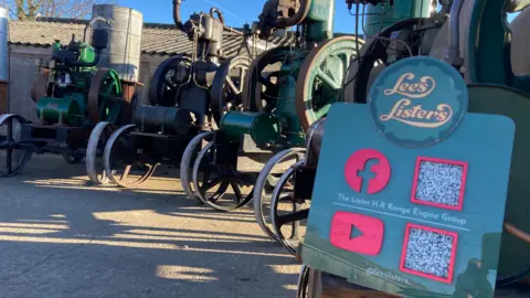 BBC/Cathy Killick A plaque reading "Lee's Listers" with the Facebook and YouTube logos, on the front of one of a number of vintage engines lined up.