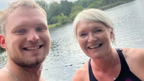 Archie Vokes, a young man with a beard wearing a silver chain around his neck, smiles for a photograph with his mother Kate. The pair are smiling for a photograph in front of what appears to be a lake. 