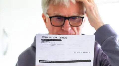 A man wearing a grey jumper and glasses is looking ay a council tax bill holding his head. He looks concerned,