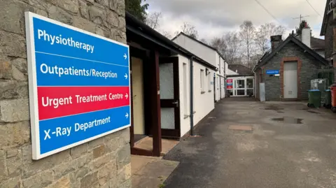 The hospital buildings are a mix of stone and white painted single-storey and two-storey buildings. A blue and red NHS sign on the exterior wall reads: "Physiotherapy. Outpatients/reception. Urgent Treatment Centre. X-Ray Department".