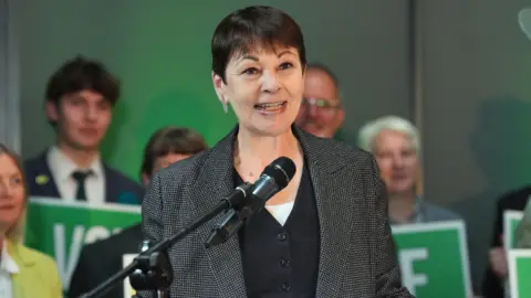 PA Media Caroline Lucas stands behind a microphone. She has a tweed-like jacket on top of a dark grey cardigan. She has short brown hair and stands in front of people holding green placards.