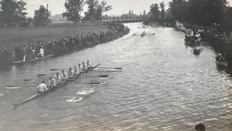 Michelle Bullivant A black and white image showing rowers competing on the River Cam. Crowds of people are gathered on the river side. Behind them can be seen grass, trees and the tall chimney of the sewage pumping station. The river bends around the corner.