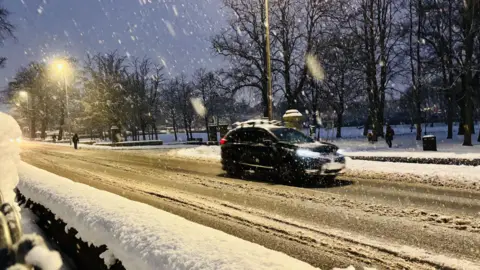 A car drives on a snow covered with melted slushy snow. Deep snow gathers at the edge of the road and it is snowing. The sky is dark blue and tall trees can be seen in the background