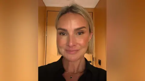 A selfie of a Katy McKinney who has short blonde hair and is smiling at the camera. She is standing in front of a wooden door and is wearing a black top and has a necklace on.