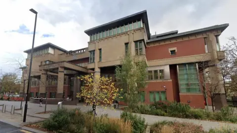 Exterior of Sheffield Crown Court, a red brick and stone building with columns and windows. In the foreground are trees and bushes.