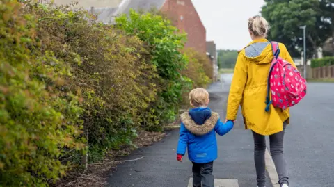 Generic picture of a boy and woman pictured from behind. The woman is wearing a yellow coat and carrying a school bag on her shoulder. The boy had a blue coat and grey trousers and he holding hands with the woman. 