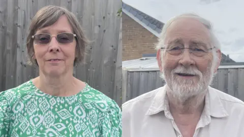 Pauline Palmer smiles at the camera, standing in front of a wooden garden fence. She has slightly darkened, square glasses on and has short light brown hair. She is wearing a green and white patterned dress. Steve Palmer has white hair and bear and is wearing a white shirt with the collar open. He is smiling at the camera.