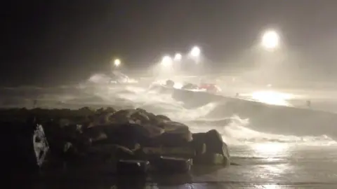 PA Media High winds causing massive waves in County Mayo, Republic of Ireland. The image is dark with bright lights glowing in the darkness amid the rain and winds.