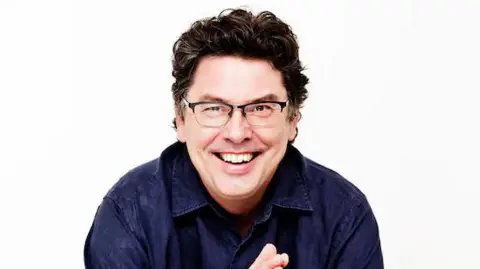 A man with brown curly hair, wearing glasses and a dark blue shirt, smiling at the camera