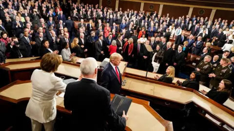 Getty Images President Donald Trump, in profile, walks to the lectern to deliver the State of the Union in 2020. Nancy Pelosi, in a white pantsuit, and former Vice-President Mike Pence are seen from behind. A full House of Representatives is in the background. 