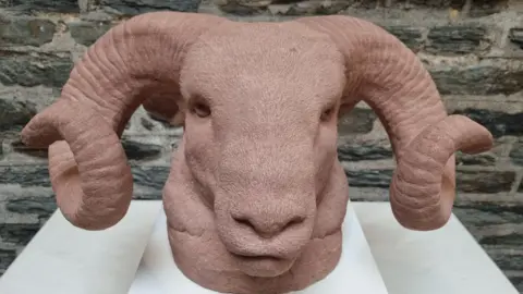 LOUISA THOMAS A close-up of a Loughtan sheep's head, sculpted in red clay, which has large spiralling horns. It is on a white plinth in front of a stone wall.
