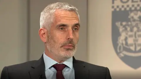 ITN A man wearing a suit and tie looking slightly off camera. He has short grey hair and a grey beard. There is part of a police symbol in the background.