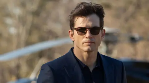 Disney via Getty Images Foto do ator Ashton Kutcher com óculos escuros, camisa preta e paletó