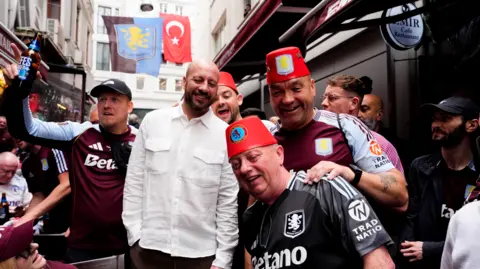 PA Media Aston Villa fans pictured in blue and claret football shirts, smiling with their arms around one another while Alan Hutton stands in the middle of the huddle, smiling wearing a white shirt.