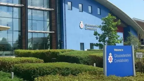 The exterior entrance of Gloucestershire Constabulary's headquarters. It is a large blue building with an arched roof and lots of windows on the left. There are manicured hedges outside the front, and a blue sign with the Gloucestershire Police logo on it. 