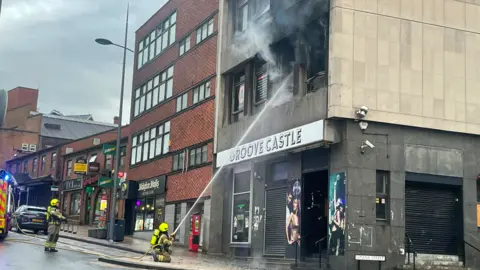 Two firefighters in protective clothing, one of them aiming a water jet at a tall grey building which appears to have been damaged by fire