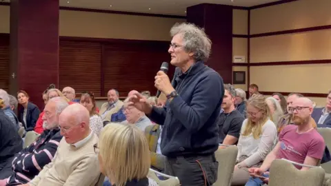 BBC Michael Kidman is wearing a blue cardigan and grey trousers. He has grey curly hair and glasses. He is speaking in a big conference room filled with people sitting down. 