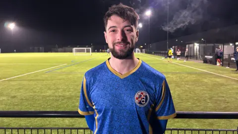 A man in a blue football shirt with a golden v-neck collar is standing looking straight at the camera with a floodlit football pitch in the background.