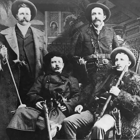 Getty Images A posed photo of four men. Cole and Bob Younger are standing at the rear, Jesse and Frank James are in the foreground. All are heavily armed and wearing large hats and long coats. 