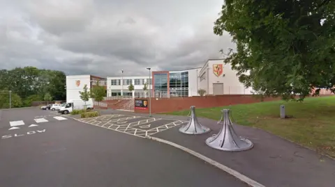 Google The front of St Margaret Ward Catholic Academy in Stoke-on-Trent, with a car park to one side and a tree in the foreground. There is a red brick wall and steps in front of the building. The building is mostly white, with large windows and a school crest on the side.