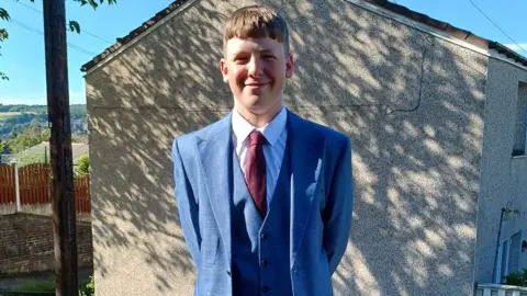 Family handout Joshua stands outdoors on a sunny day. He is dressed in a formal outfit consisting of a light blue three-piece suit, a white dress shirt, and a dark red tie. Behind him, there is a beige-coloured building with a sloped roof and a small window visible on the side. A wooden fence and some trees can be seen in the background, along with a clear blue sky and distant hills.