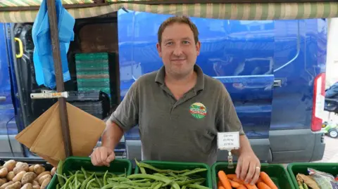 A man with short brown hair, wearing a grey polo shirt, stands behind his fruit and vegetable stall at a market, with green beans and orange carrots laid out in front of him. A blue van is parked in the background.