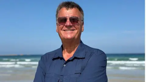 Graeme Lowe A picture of author Graeme Lowe wearing a blue shirt and dark brown sun glasses on. He is stood on a beach on a sunny day smiling at the camera. 