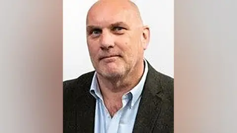 Hampshire County Council A head and shoulder image of Councillor Nick Adams-King, who has a bald head, is clean shaven and is wearing a light blue shirt with the collar undone and a dark suit jacket