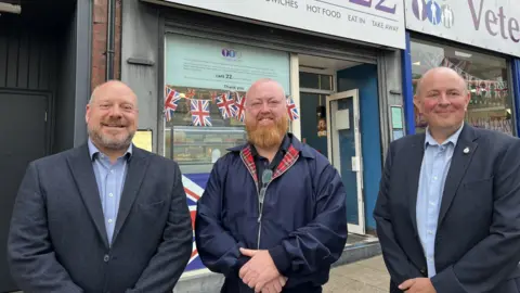 Three men standing in the street in front of a cafe and Veterans at Ease shop. They are smiling at the camera. Garreth, on the left, has short, light brown hair, and stubble; he is wearing a navy blue jacket and light blue shirt. Mark, in the centre, is bald with a ginger beard, wearing a blue jacket with a tartan lining. Andrew, on the right, has fine light hair and wears a navy jacket and light blue shirt.