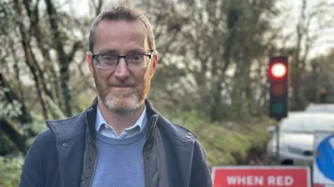 Philip McGuigan MLA is wearing a blue jumper with a blue shirt underneath. He is wearing a navy jacket and glasses. Philip is smiling at the camera and has red and grey stubble and short hair.