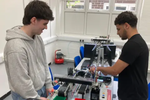 William Taylor and Saleh Mansour at Neath College. Saleh is on the right in a black T-shirt, while William is on the left in a grey hoody. Both are stood up and are working on an engineering project, which is in the middle of them.