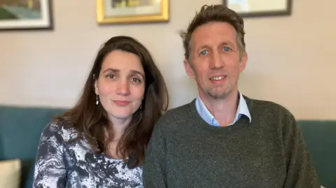 Andrew Davies (right) and his wife Milica. They are sitting on a green sofa and pictured from the chest up. She has long dark hair, is wearing a black and white marble-effect dress and drop pearl earrings. He has on a blue shirt with the collar unbuttoned and a dark green jumper over the top.