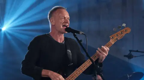 A wide shot of Sting playing a bass guitar. He has short grey hair and is wearing a black jumper.
