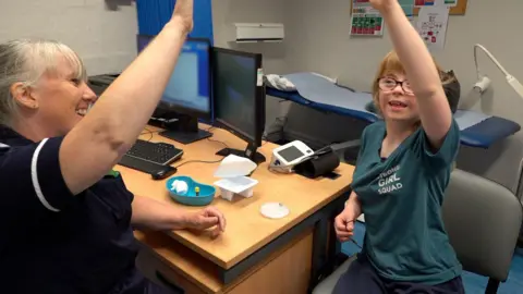 A nurse raises her hand in the air to give a 'high-five' to a young girl. The pair are sat together in a nurse's office.