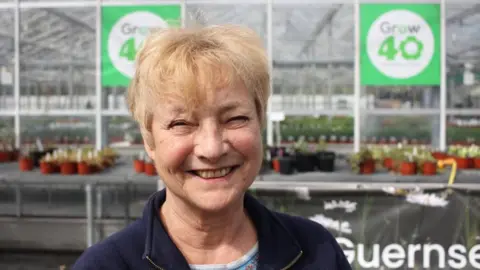 A woman with short blonde hair smiles at the camera in a navy jacket with a plant greenhouse in the background