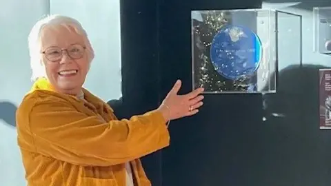 Eric Morecambe Centre Gail Morecambe wearing a mustard yellow coat and white hair is gesturing to a blue circle plaque in a clear plastic box. She is smiling at the camera. 