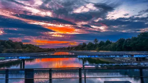 Lewis Jefferies A bright orange sunset lights up the sky with vivid colours. The light is reflected in the water below making it glow a bright orange colour. The water has a grey bridge over it and the banks are flanked by trees.